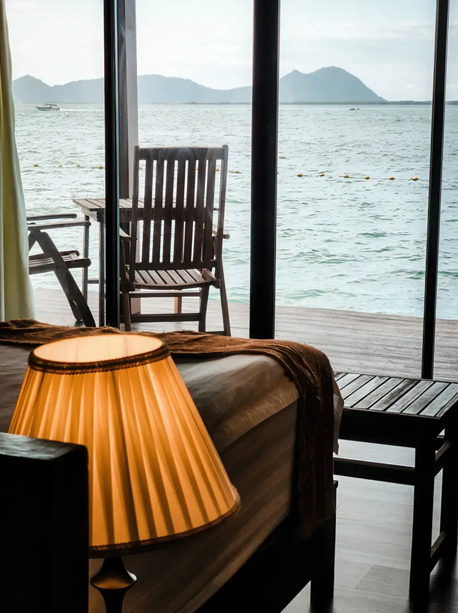 A cozy bedroom at Sea Star Resort in the Celebes Sea displaying a bed facing panoramic glass doors opening onto the balcony, a bedside lamp, rich mahogany style wooden floors, and aircon.